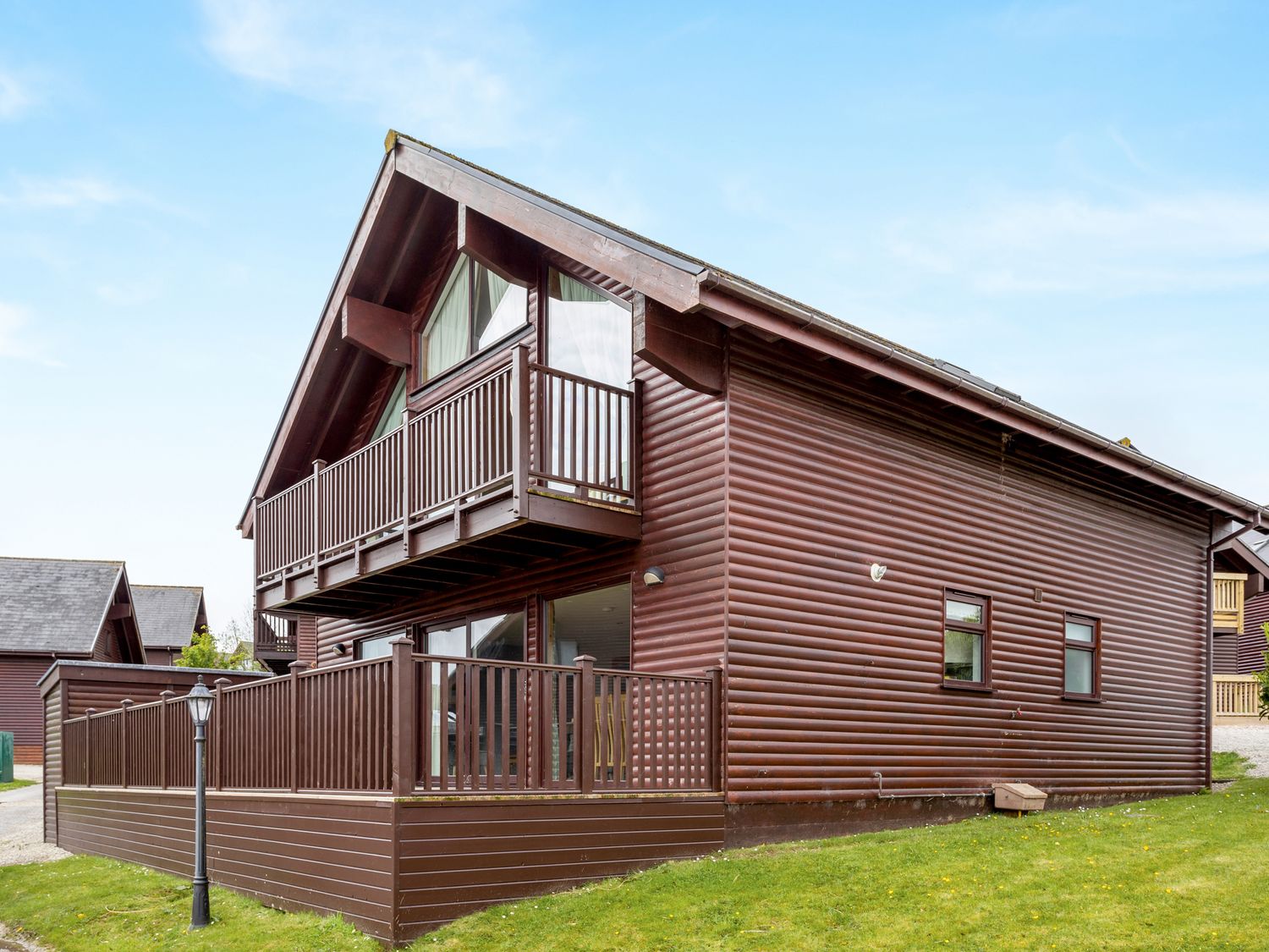 A wooden cabin with a balcony and fenced patio on a grass lawn at Sunbow at Retallack Resort in Winnards Perch St Columb Major