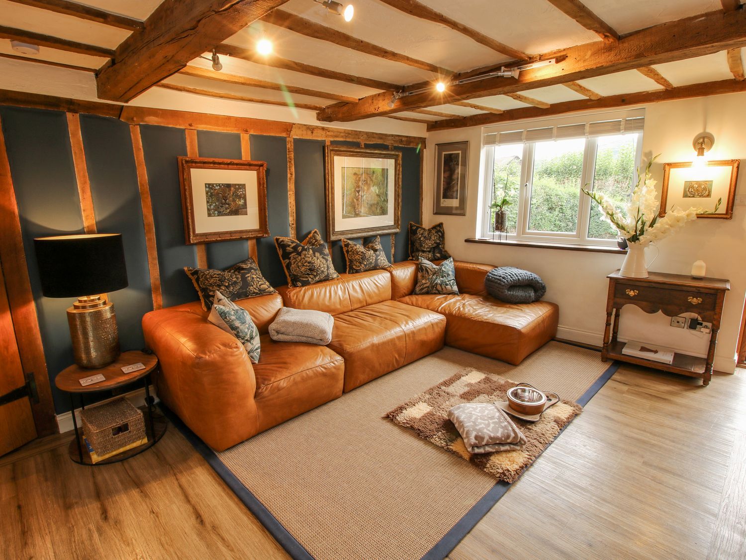 A living room with a tan leather sectional sofa and cushions a wooden side table with a lamp and framed pictures on the wall at Pen Y Groes in Berriew