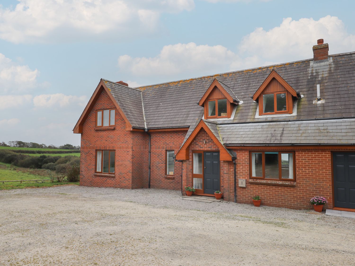 A brick house with a gray tiled roof and multiple windows surrounded by a gravel driveway and green fields at Halls Farm in Poughill near Bude