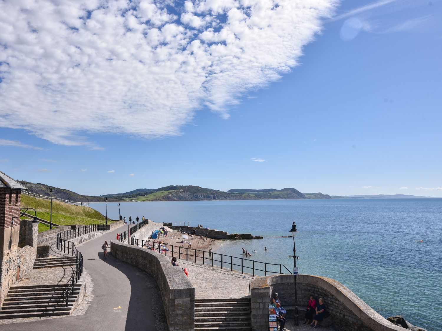 A seaside walkway with people walking and sitting near the water and a rocky beach at Marnies Rest in Lyme Regis