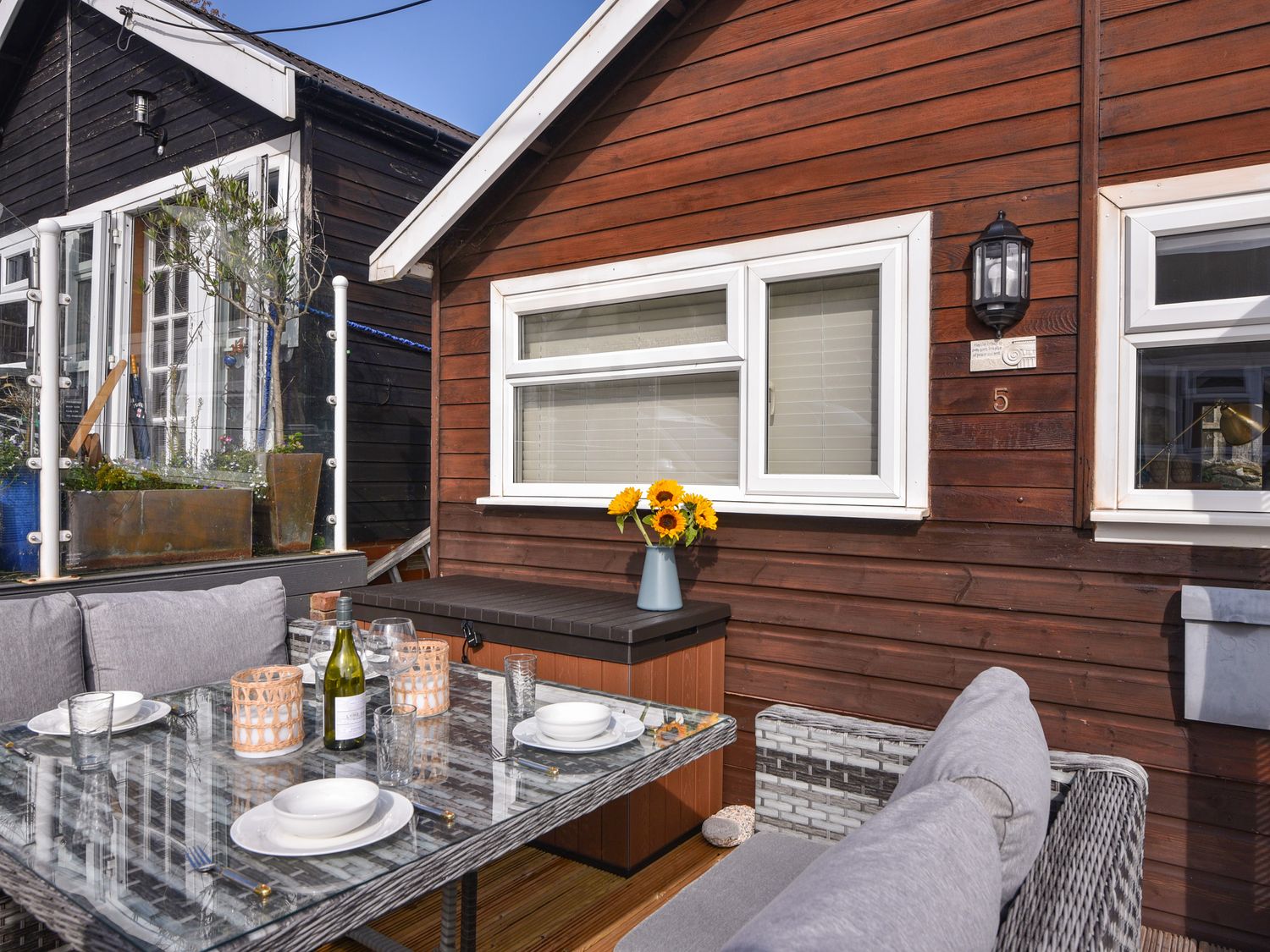 An outdoor dining area with a glass table set with plates glasses and candles beside a wooden building with windows at Marnies Rest in Lyme Regis