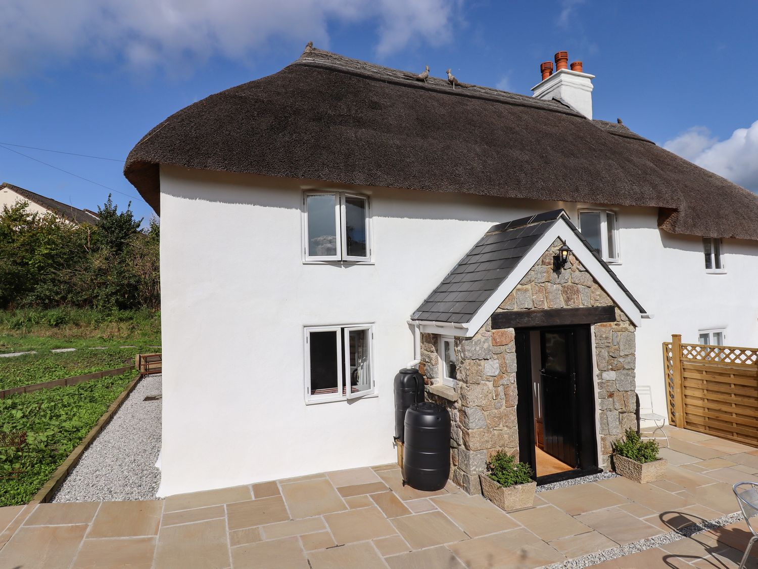 A white cottage with a thatched roof a stone porch and two black water barrels outside at Cob Cottage in Bovey Tracey