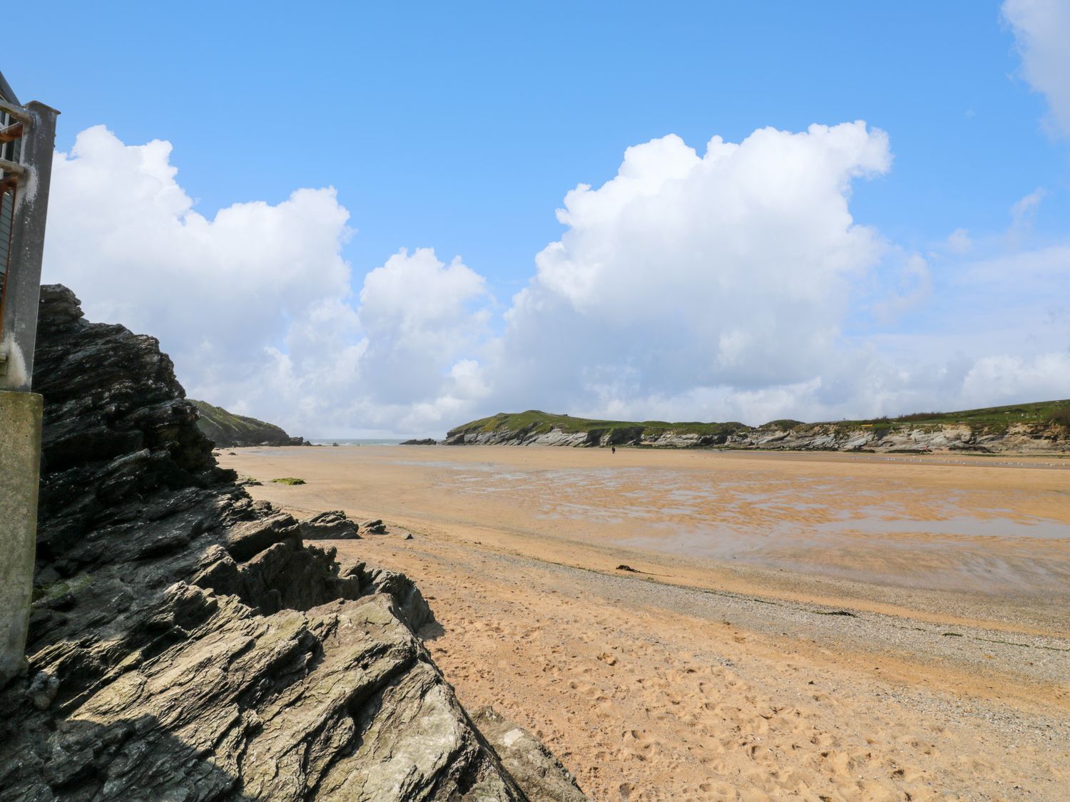 A sandy beach with rocky formations on the left and grassy cliffs in the distance under a blue sky with clouds at 119 Henver Road in St Columb Minor