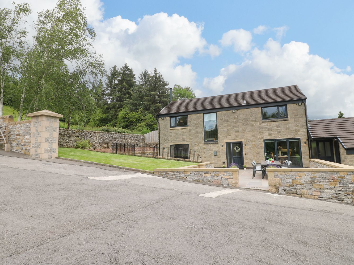A stone house with a tiled roof a patio area with a table and chairs and a lawn with trees at Ty Gambo in Oker near Matlock