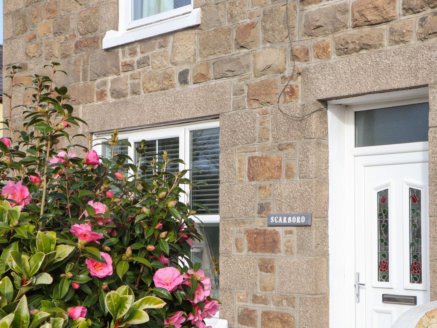 A stone exterior wall with a white door and window and pink flowers in front at Scarboro in Redruth