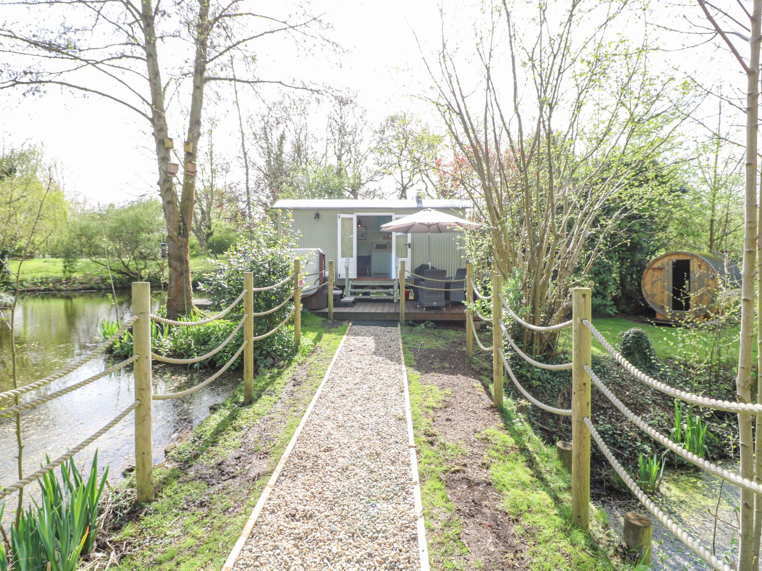A gravel pathway with rope fencing leading to a small hut with outdoor seating and an umbrella next to a pond at Damsel Shepherds Hut in Badwell Ash