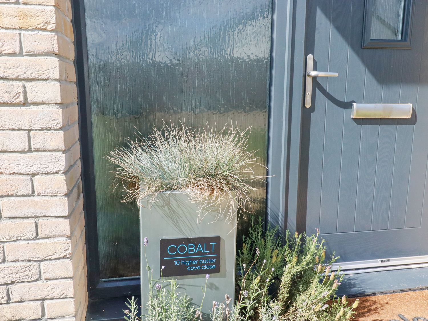 A door with a silver handle and letterbox next to a planter with grass and a sign saying cobalt 10 higher butter cove close at Cobalt in St Eval near Mawgan Porth