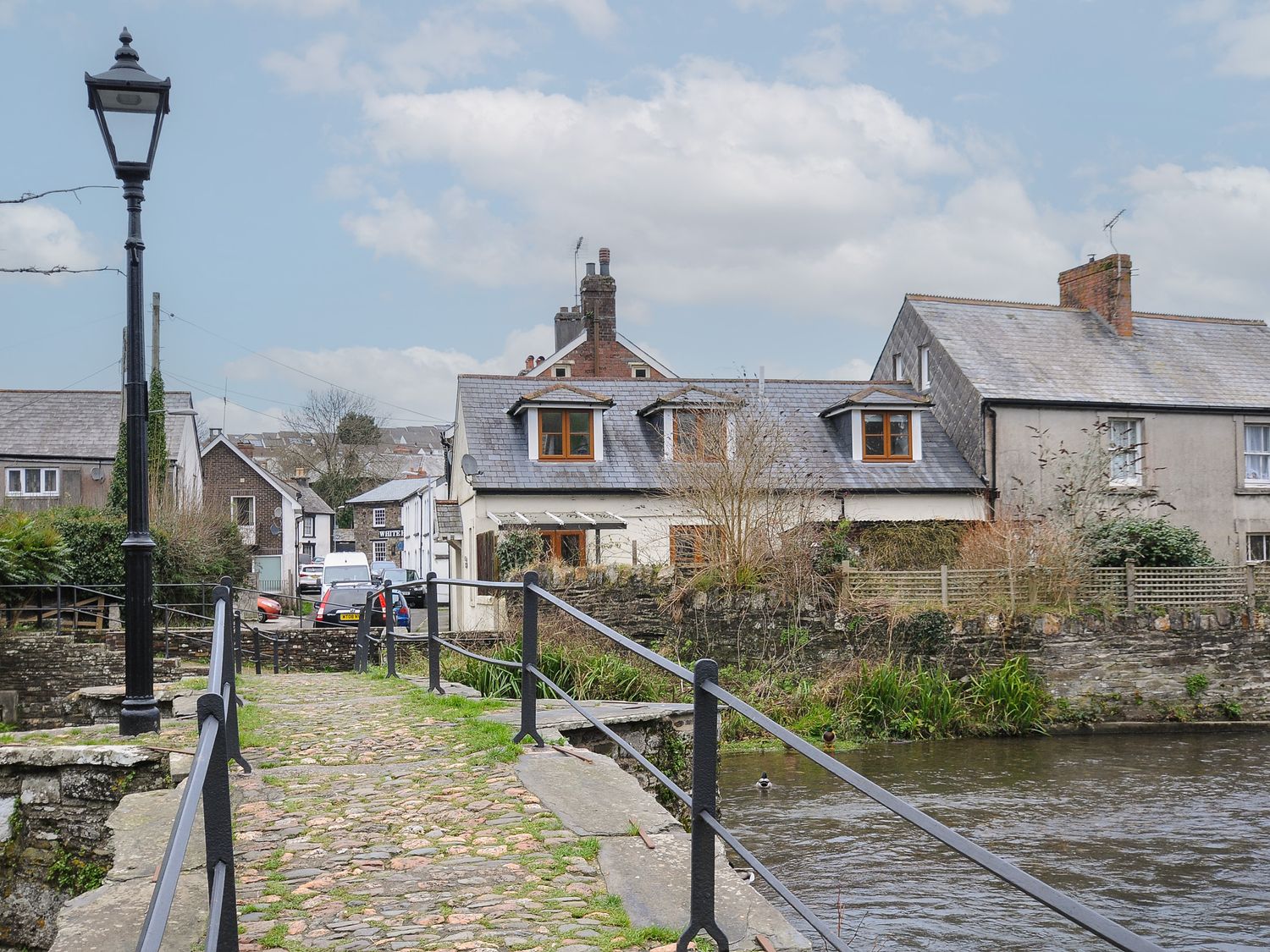 A stone footbridge over a river with houses in the background and a lamp post at Rainbows End in Launceston