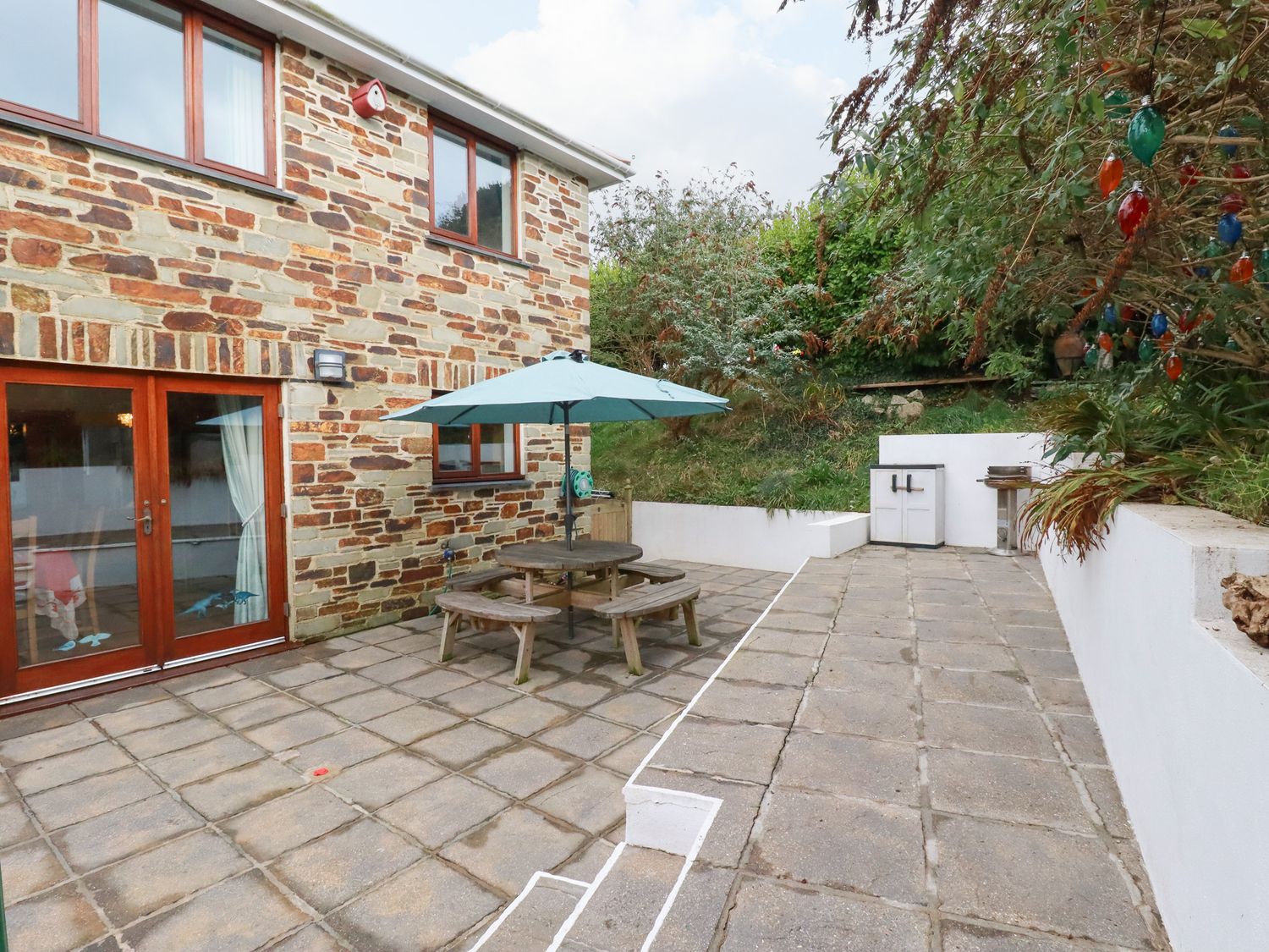 An outdoor patio area with stone paving a wooden picnic table with umbrella and a barbecue grill at Lowarth Chi Polruan