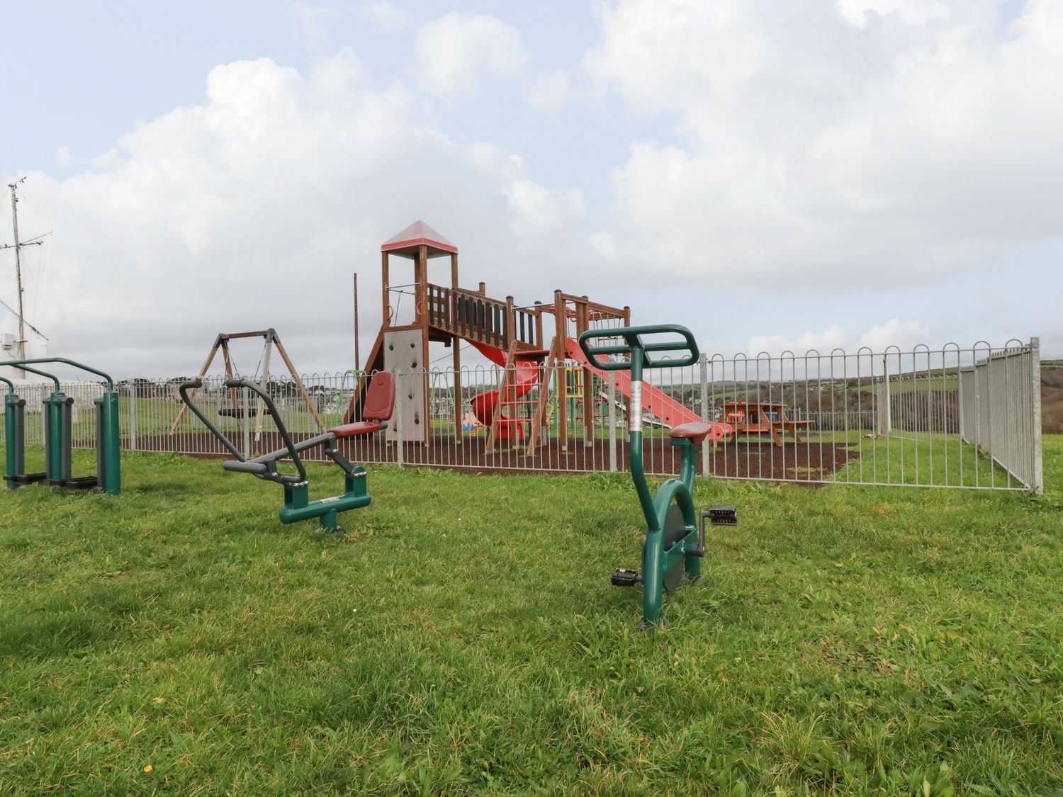 An outdoor playground with a slide tower a swing set fenced area and exercise equipment on grass at Lowarth Chi in Polruan
