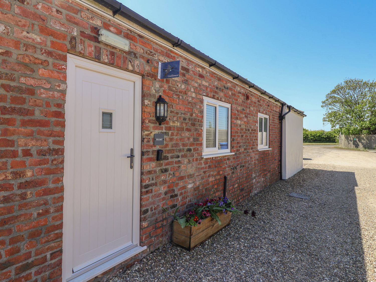 Exterior view of a brick building with a white door two windows and a flower box on gravel at Guillemot Flamborough