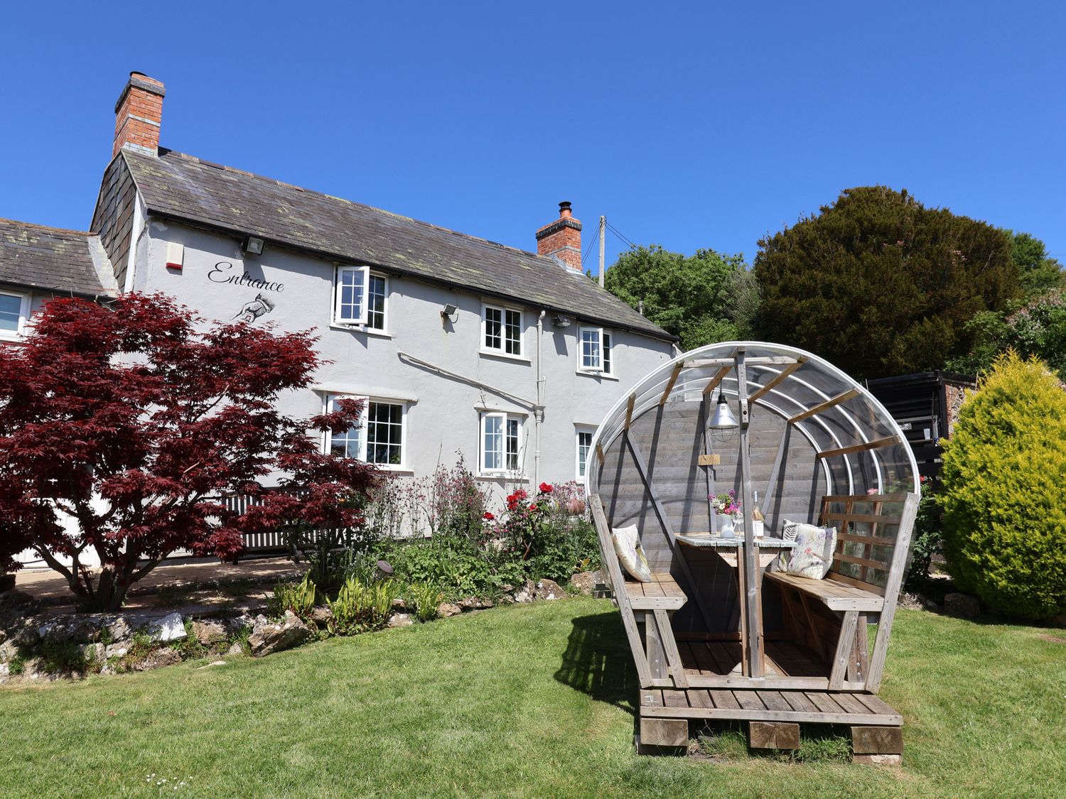 A garden with a white house in the background and a wooden covered seating area in the grass at Eggardon Rest in Askerswell near Bridport
