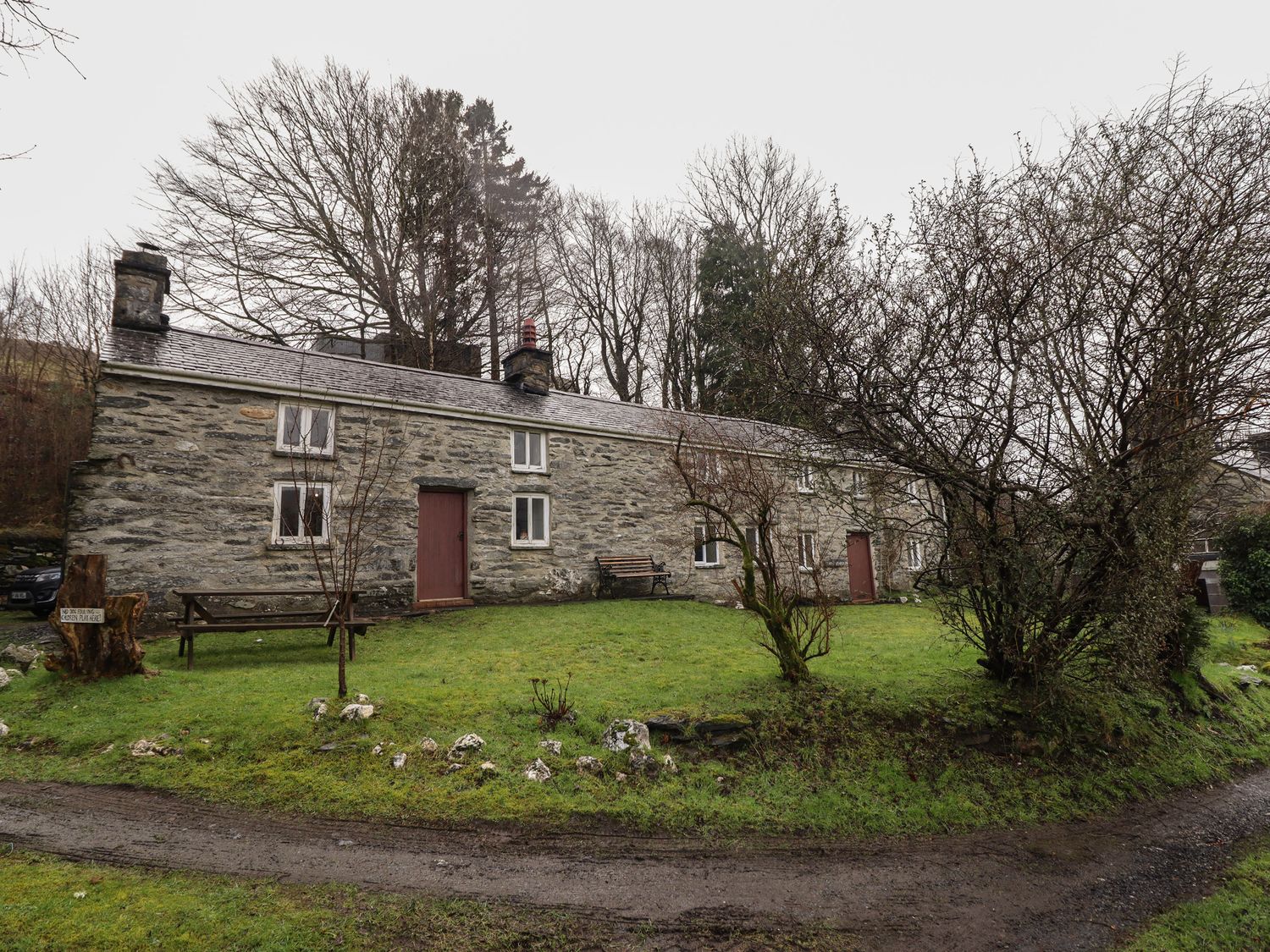 A stone cottage with red doors and white windows in a grassy yard with trees and benches at The Long House in Penmachno