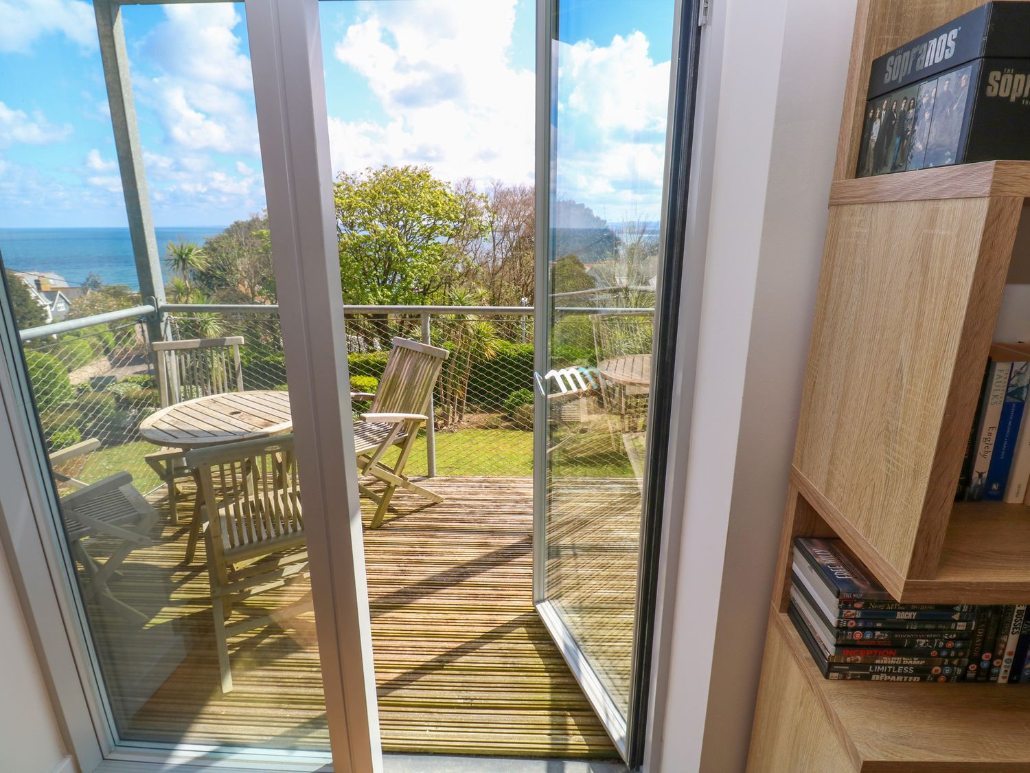 A balcony with wooden table and chairs overlooking trees and sea at Talland View in St Ives