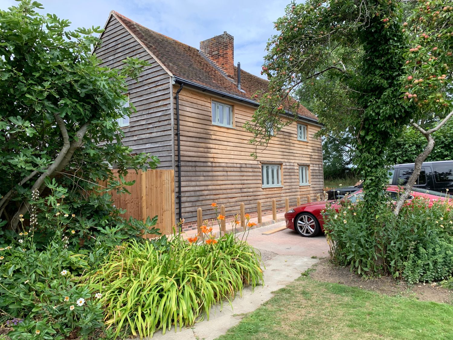 A wooden house with a tiled roof next to a garden with trees flowers and a red car in a driveway at Lodge Cottage in Harwich