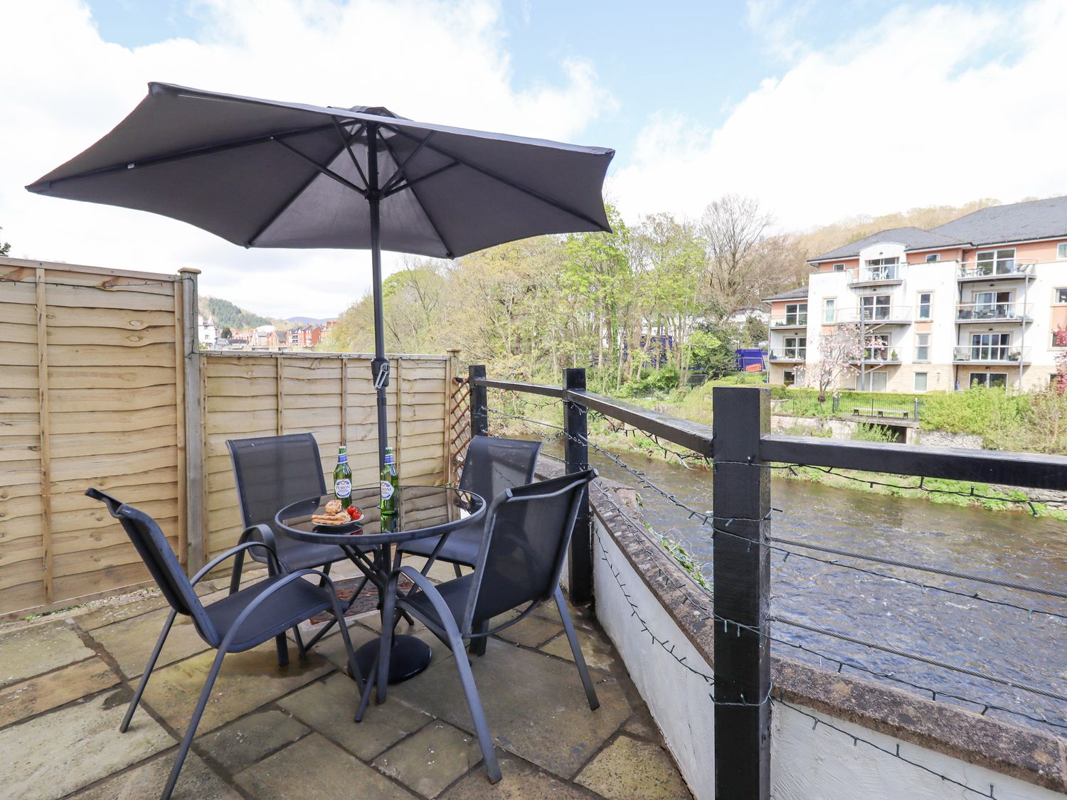 An outdoor patio with a glass table and four chairs under a large umbrella overlooking a river at No 9 On The Riverbank in Llangollen