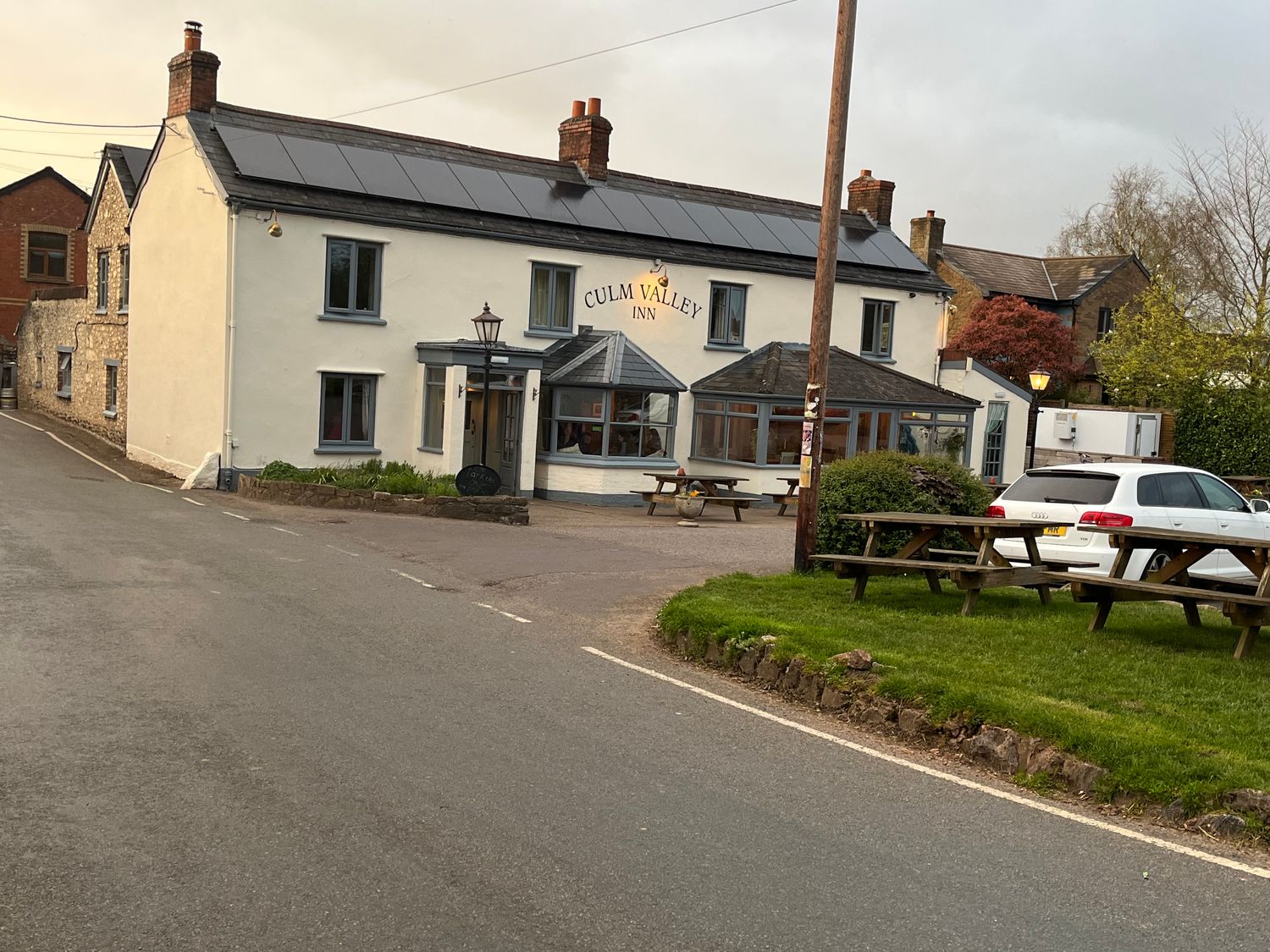 A street view of Culm Valley Inn building with parked white car and wooden picnic tables on grass at Piglet Corner in Culmstock