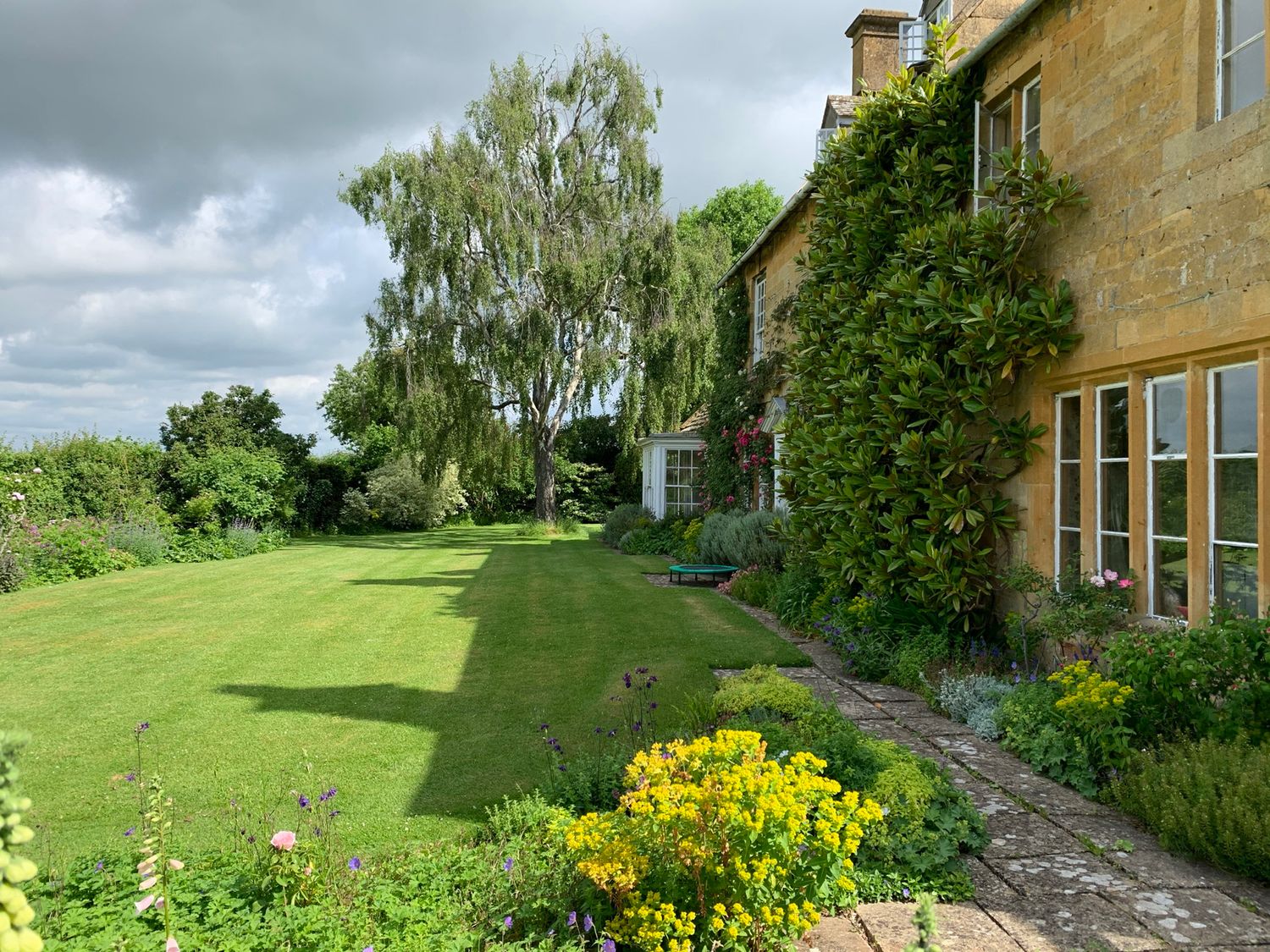 A garden with a lawn trees and flowers beside a stone house at Wood Stanway House Wood Stanway near Toddington