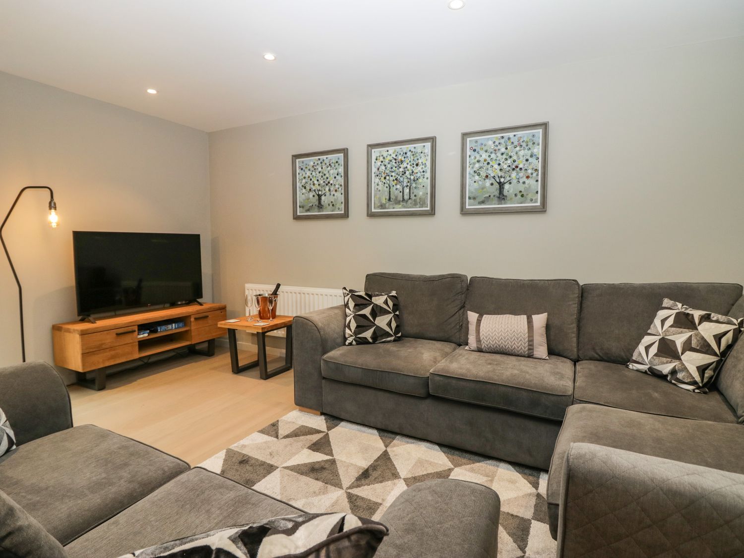 A living room with a grey sectional sofa a patterned rug a wooden TV stand with a television and three paintings of trees on the wall at Brimstone in Backbarrow near Ulverston