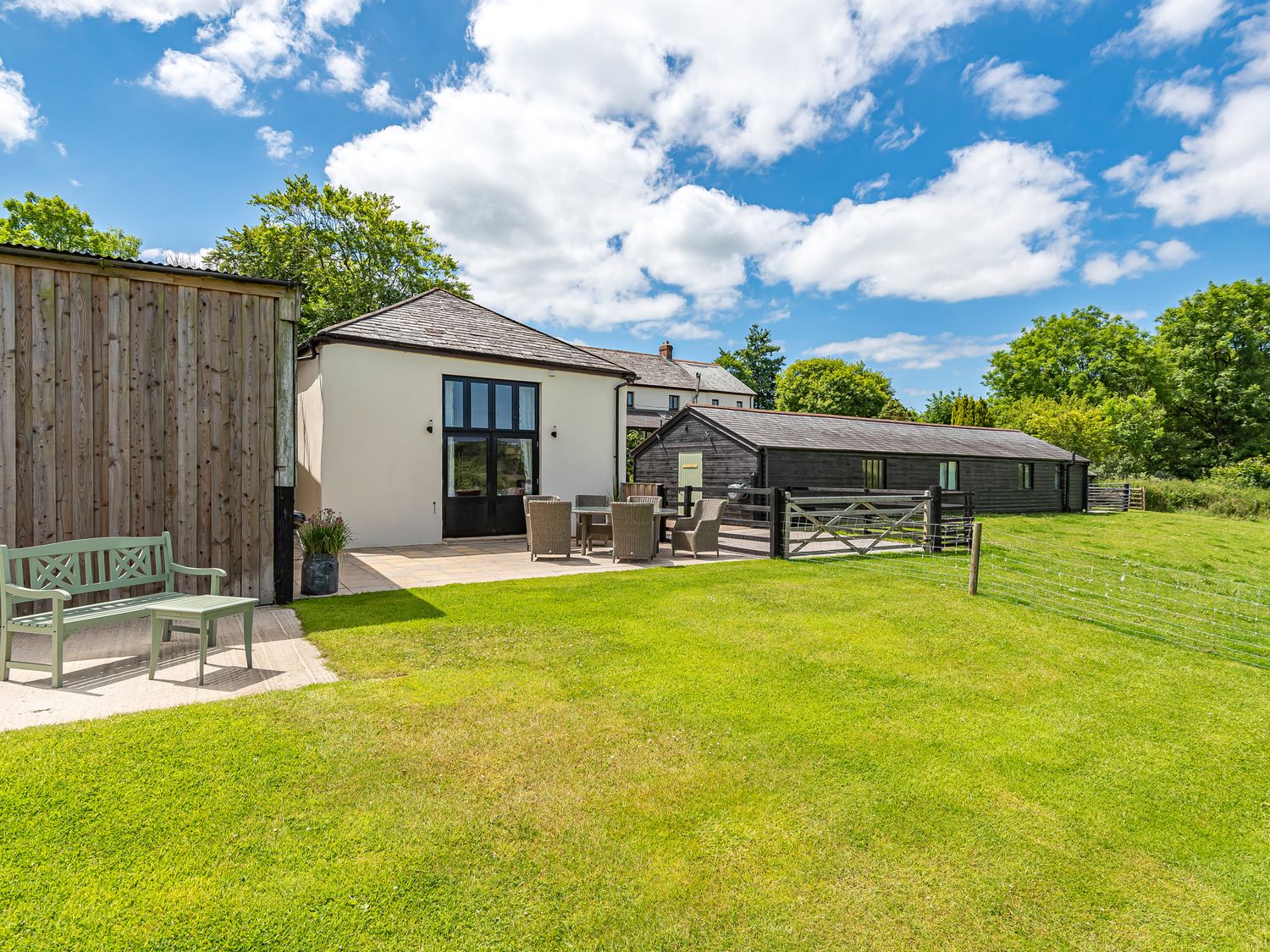A grassy yard with outdoor seating and three buildings under a blue sky at Waterland Old Barn in Bradworthy