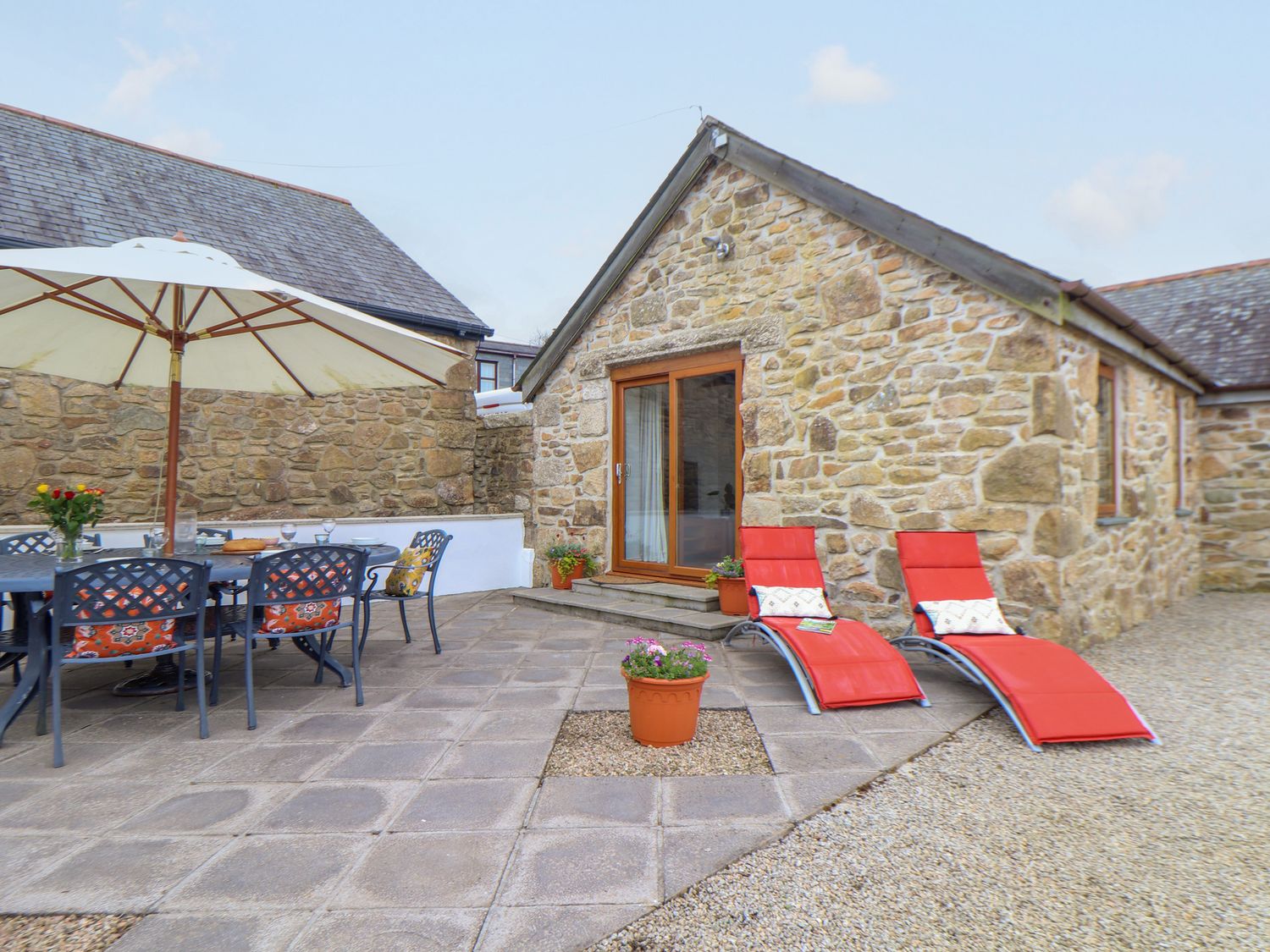 An outdoor patio with a dining table and chairs under an umbrella two red lounge chairs and a stone building at Riviere Barton in Hayle