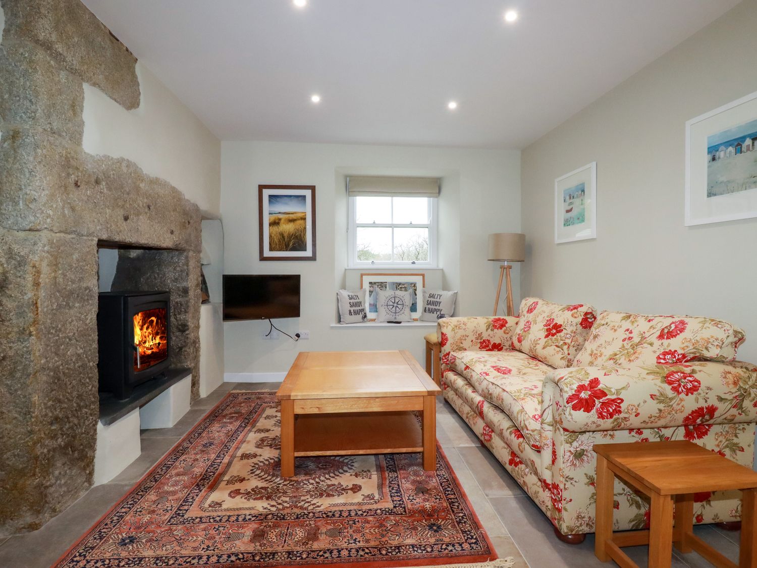 A living room with a floral sofa, wooden coffee table, stone fireplace with a burning fire, wall-mounted TV, window with cushions, and a floor lamp at Tretoil Farm House in Bodmin