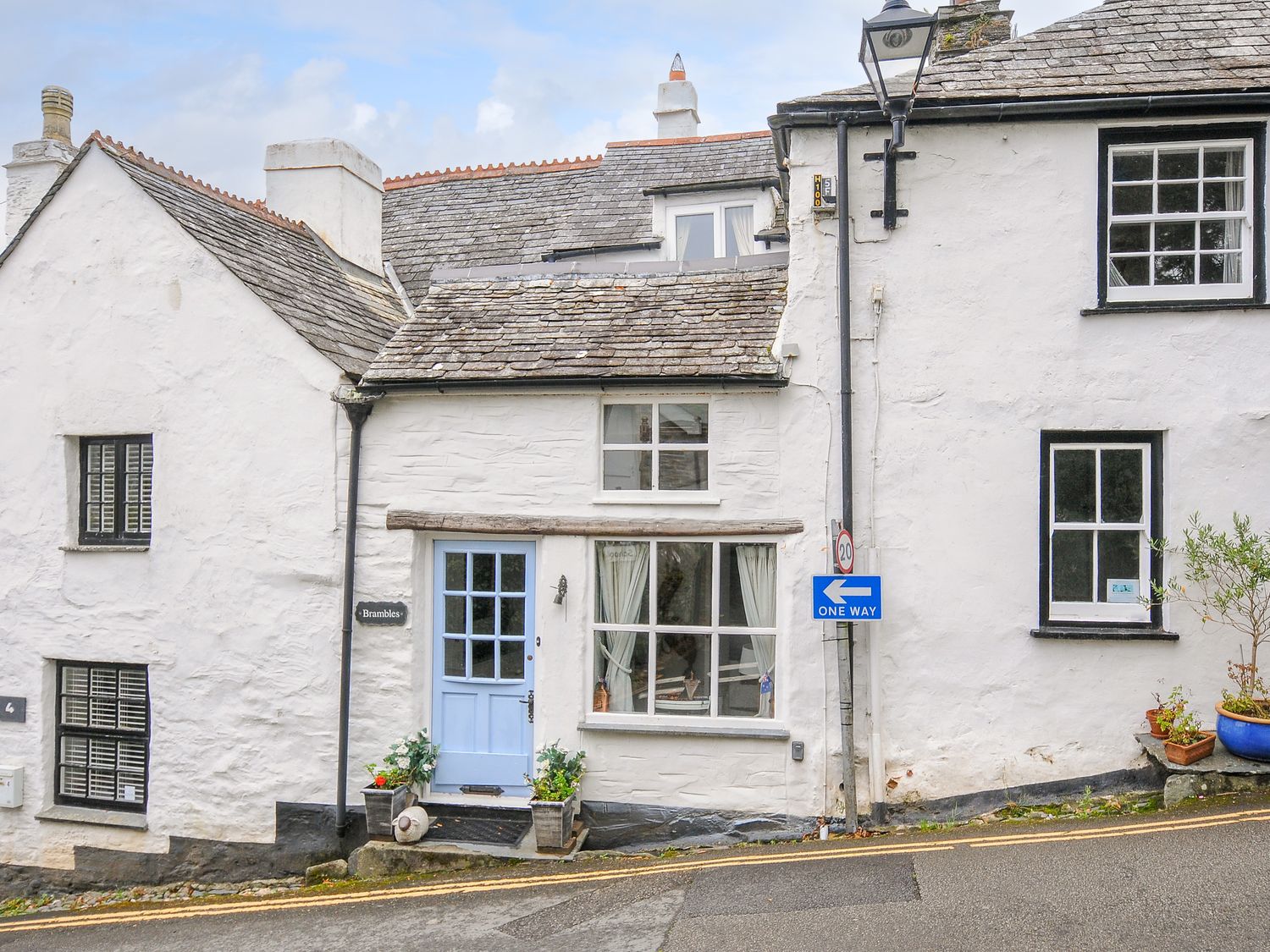A white stone cottage with a blue door and window boxes on a street with a one way sign at Brambles in Boscastle