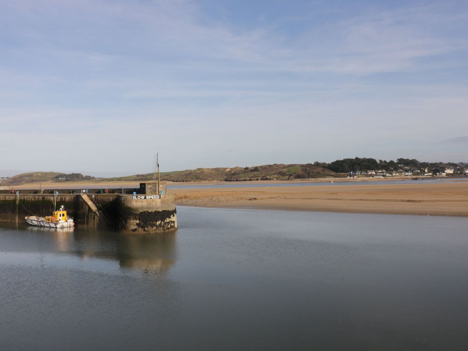 A small harbor with a dock and a boat in the water with a sandy beach and hills in the background at Polwen in Harlyn Bay Padstow