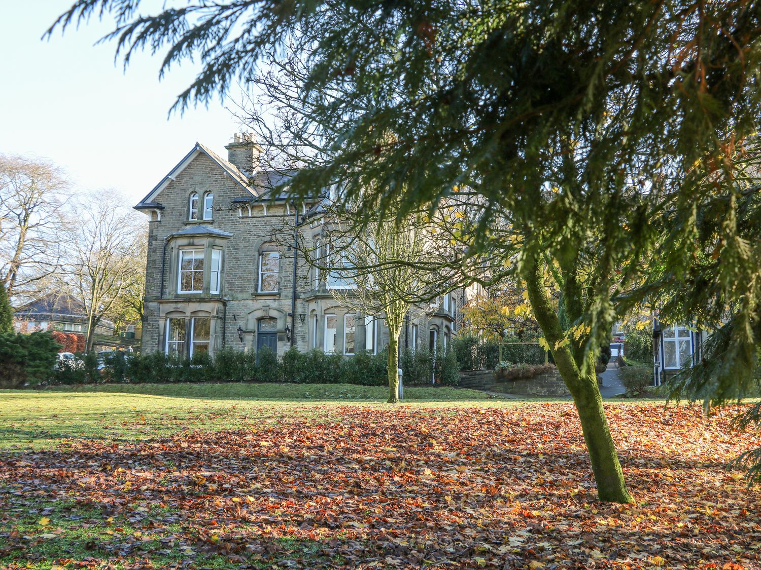 An old stone house with windows and a front door viewed from a garden with grass trees and fallen leaves at Castleton Suite in Buxton