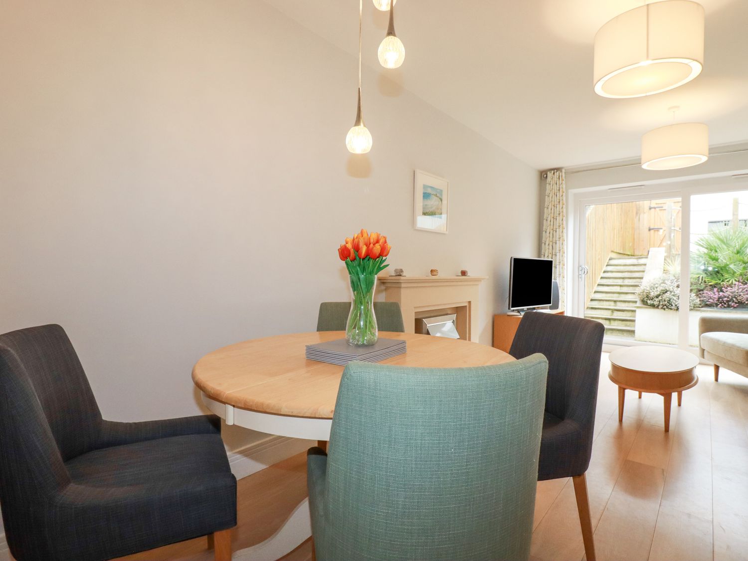 A dining area with a round table and four chairs a vase with orange flowers on the table a fireplace a television a coffee table and sliding glass doors leading outside at 17 Ocean Heights in Newquay