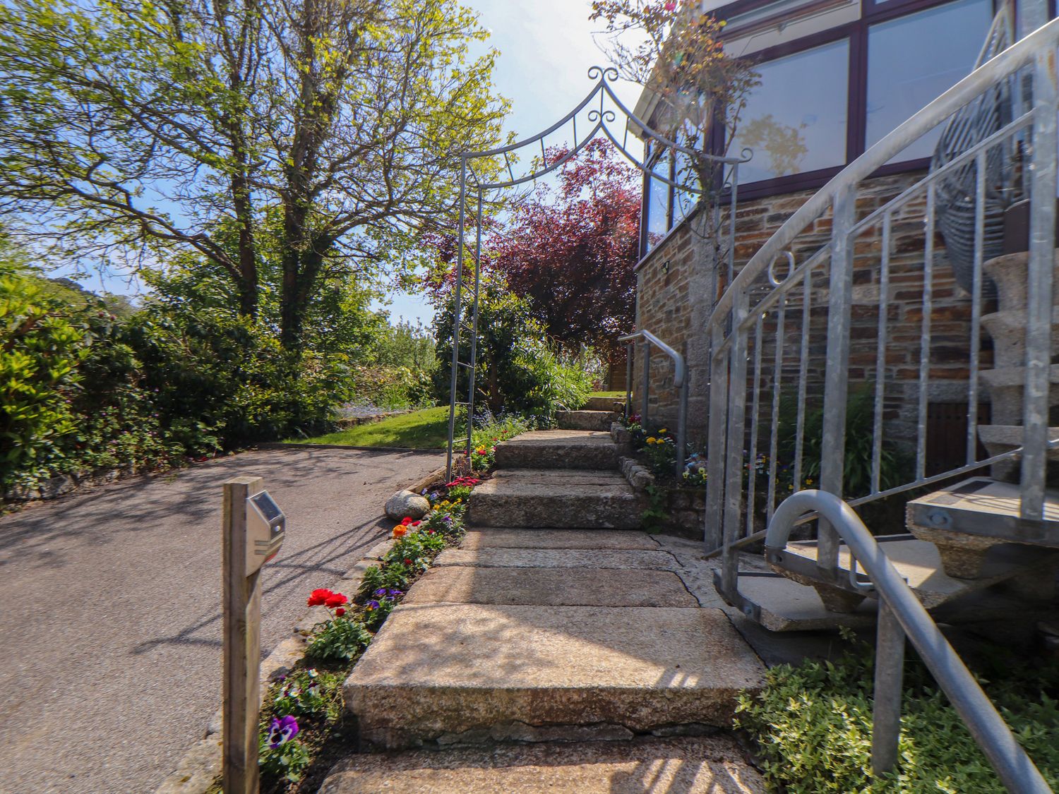 Stone steps leading up to a garden arch and a house with a spiral metal staircase surrounded by plants and flowers at Lakeside in Falmouth