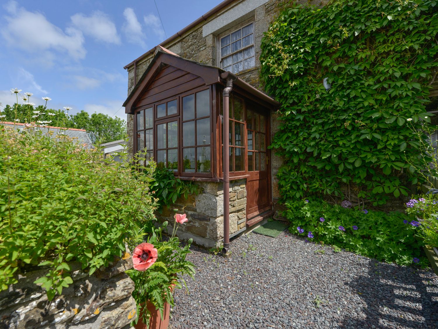 Stone house with wooden porch surrounded by plants and flowers at Swallow Cottage in Mawgan
