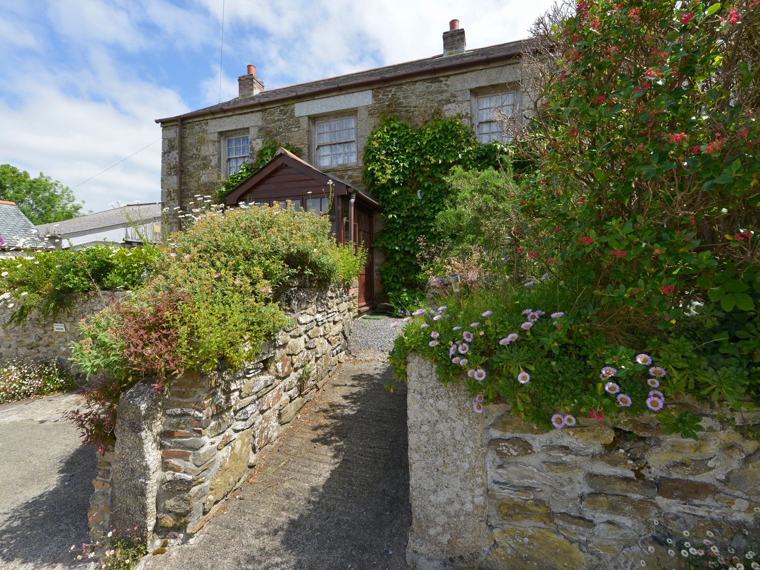 A stone cottage with a wooden entrance surrounded by flower beds and climbing plants at Swallow Cottage in Mawgan