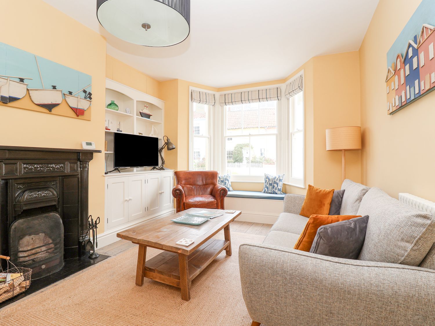 A living room with a gray sofa orange pillows a brown leather armchair wooden coffee table black fireplace and built in shelves with a TV at The Corner Cottage in Aldeburgh
