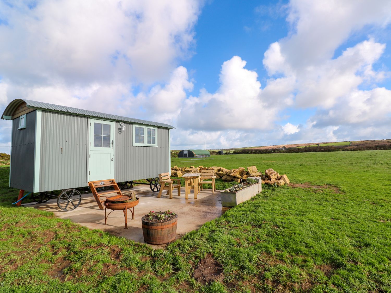 A shepherds hut on wheels with a patio area furniture and a fire pit in a grassy field at Lower Trewern Shepherds Hut in Penzance