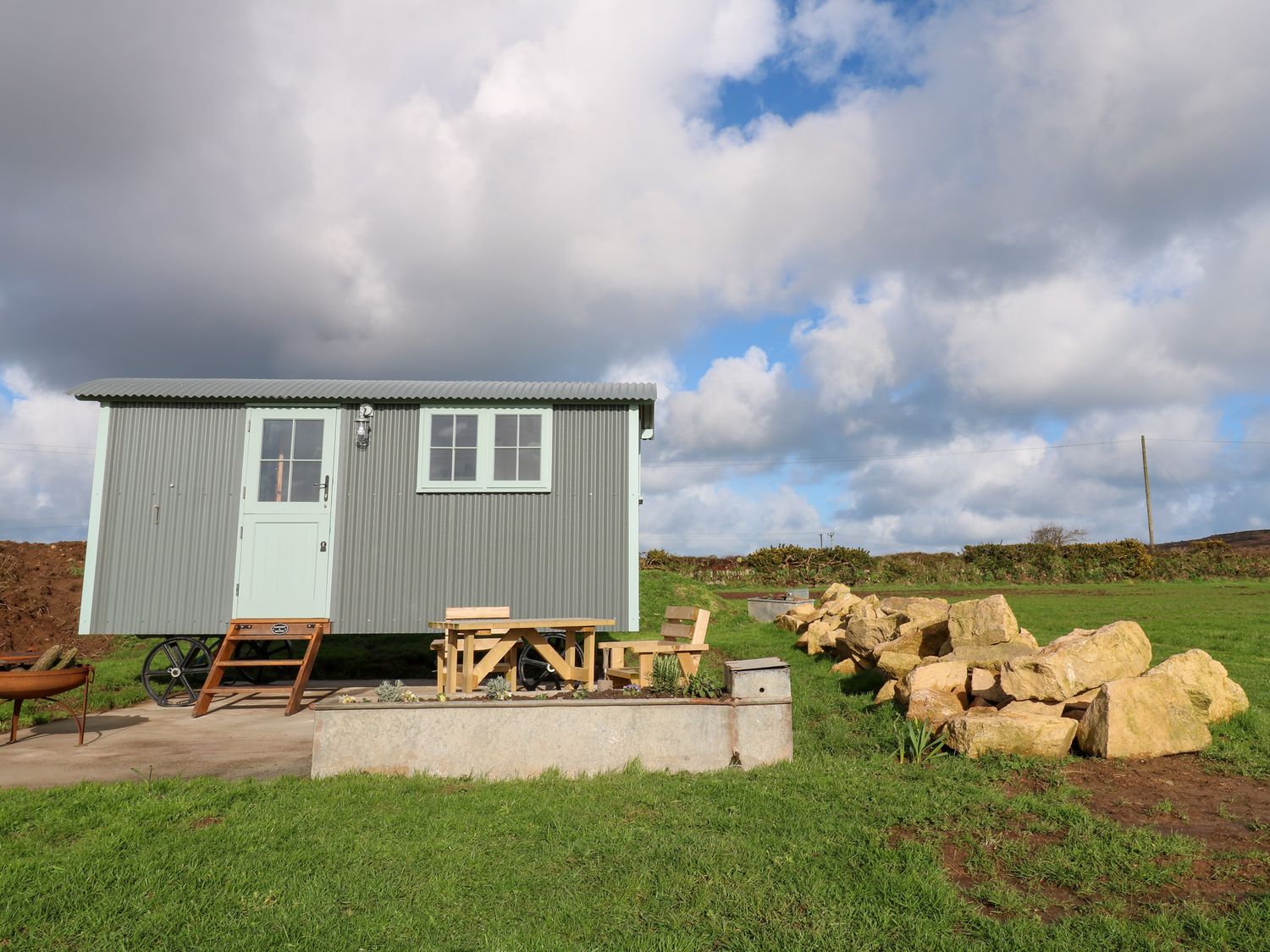 A small gray shepherds hut with a wooden bench and table outside on grass at Lower Trewern Shepherds Hut in Penzance