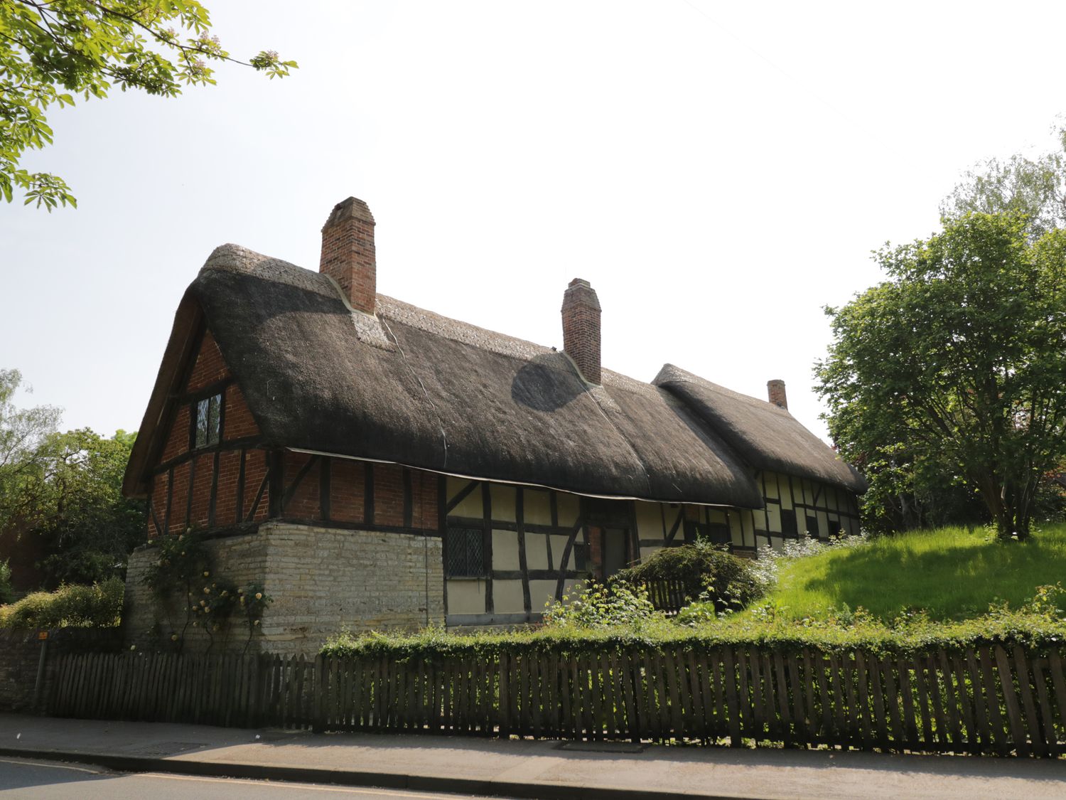 A thatched roof cottage with brick chimneys surrounded by trees and a wooden fence at Shirleys in Stratford-Upon-Avon