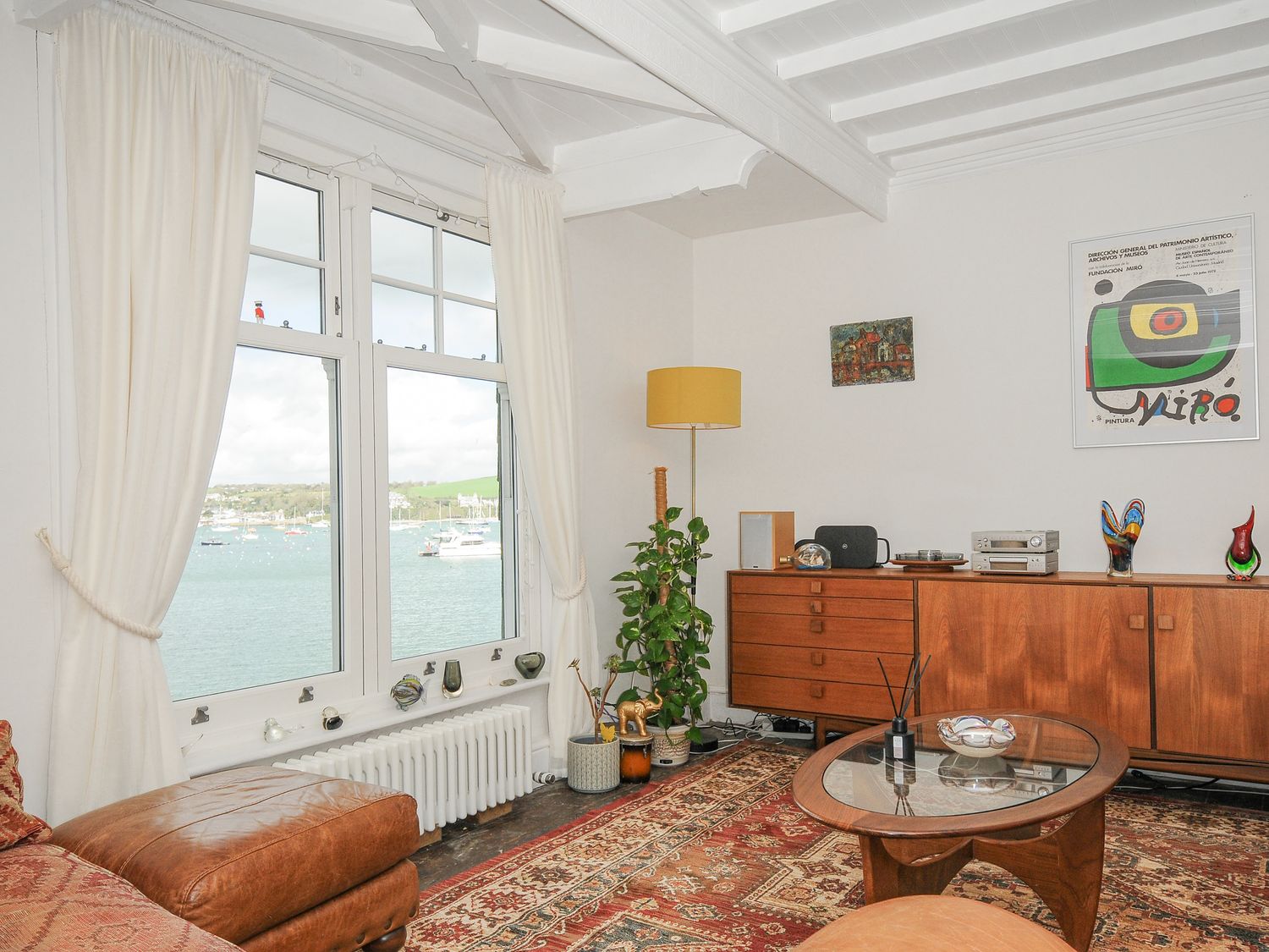 A living room with a large window overlooking water a wooden cabinet a glass coffee table and a patterned rug at Zawn Haven - Waterfront House in Falmouth