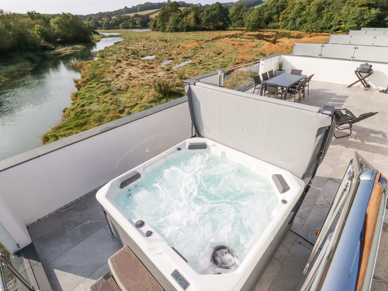 An outdoor area with a hot tub a dining table with chairs a barbecue grill and a river view at Carnon Gwel Devoran in Devoran between Falmouth and Truro