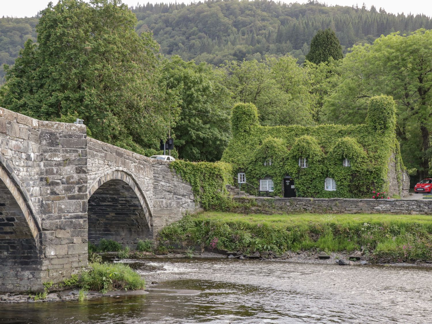 A stone bridge over a river next to a house covered in green ivy with trees and hills in the background at Royal Oak Farm Cottage in Betws-y-Coed