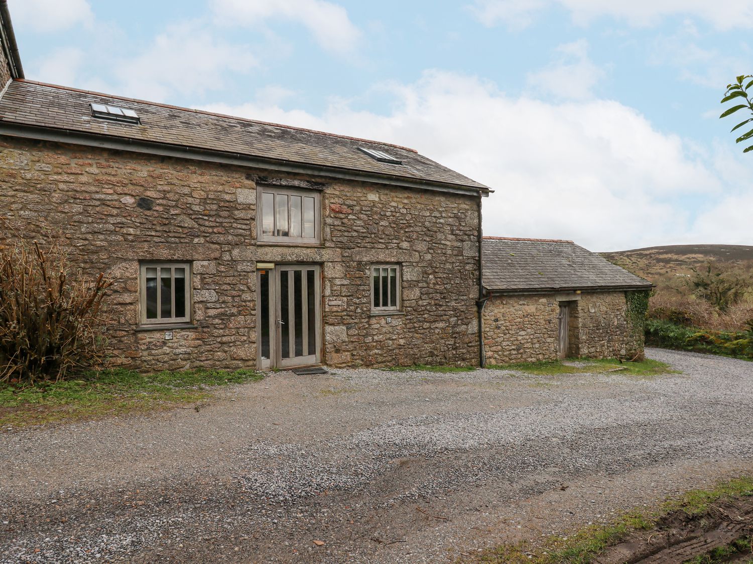 A stone barn with two windows and a double glass door surrounded by gravel and grass at Brimpts Barn Dartmeet near Princetown