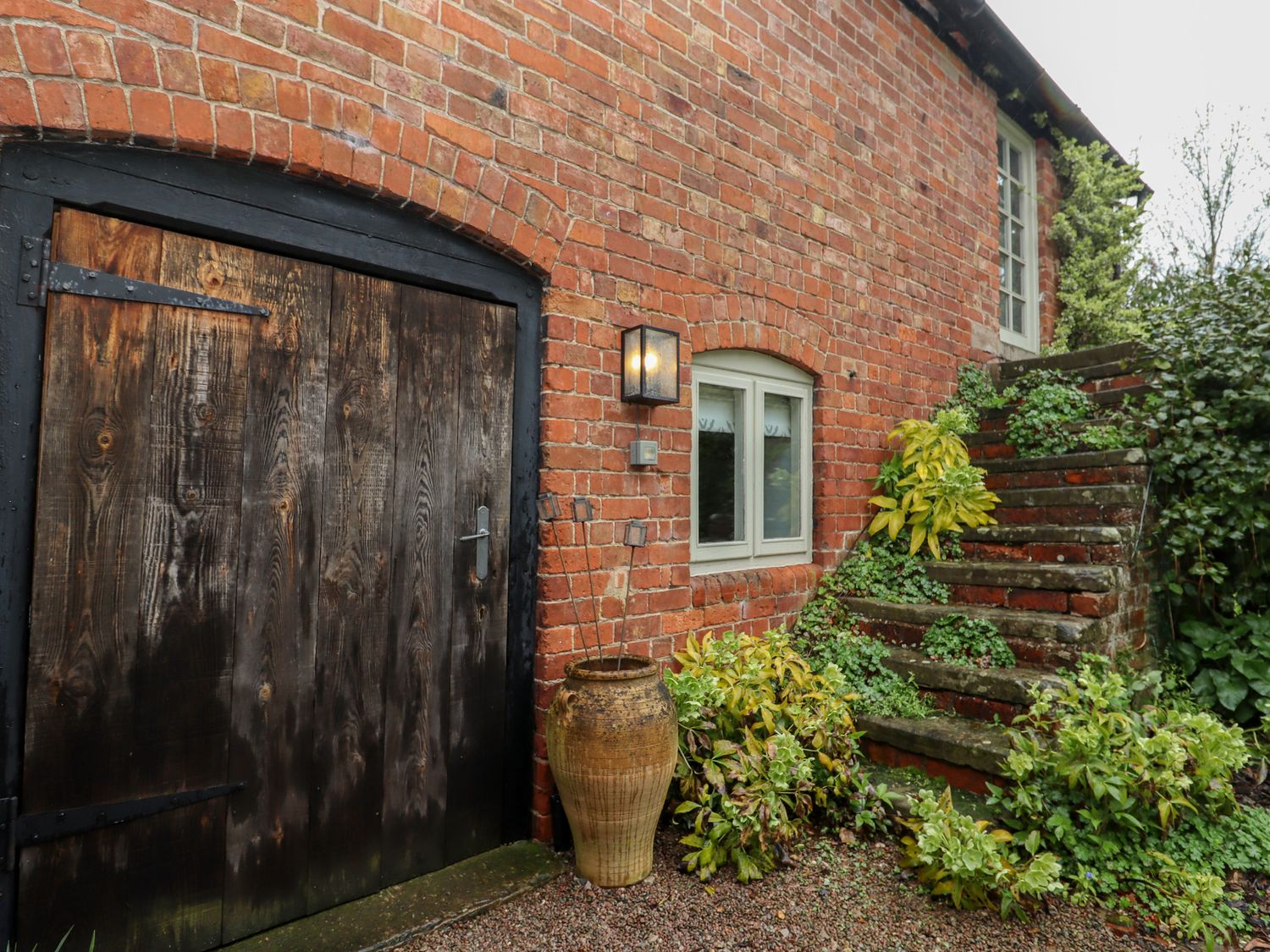 Exterior view of a brick building with a large wooden door small window stone steps and plants at Millpond Bothy in Woolhope