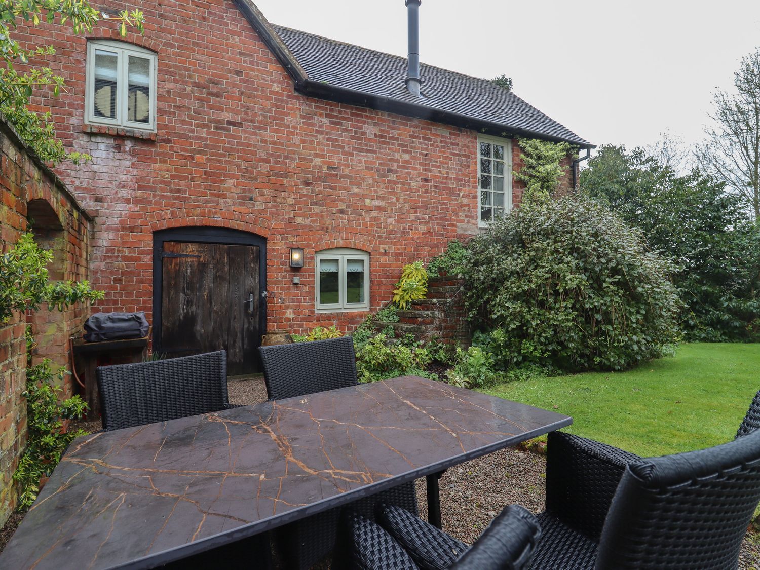 An outdoor patio with a table and chairs next to a brick house with plants and a grassy lawn at Millpond Bothy in Woolhope