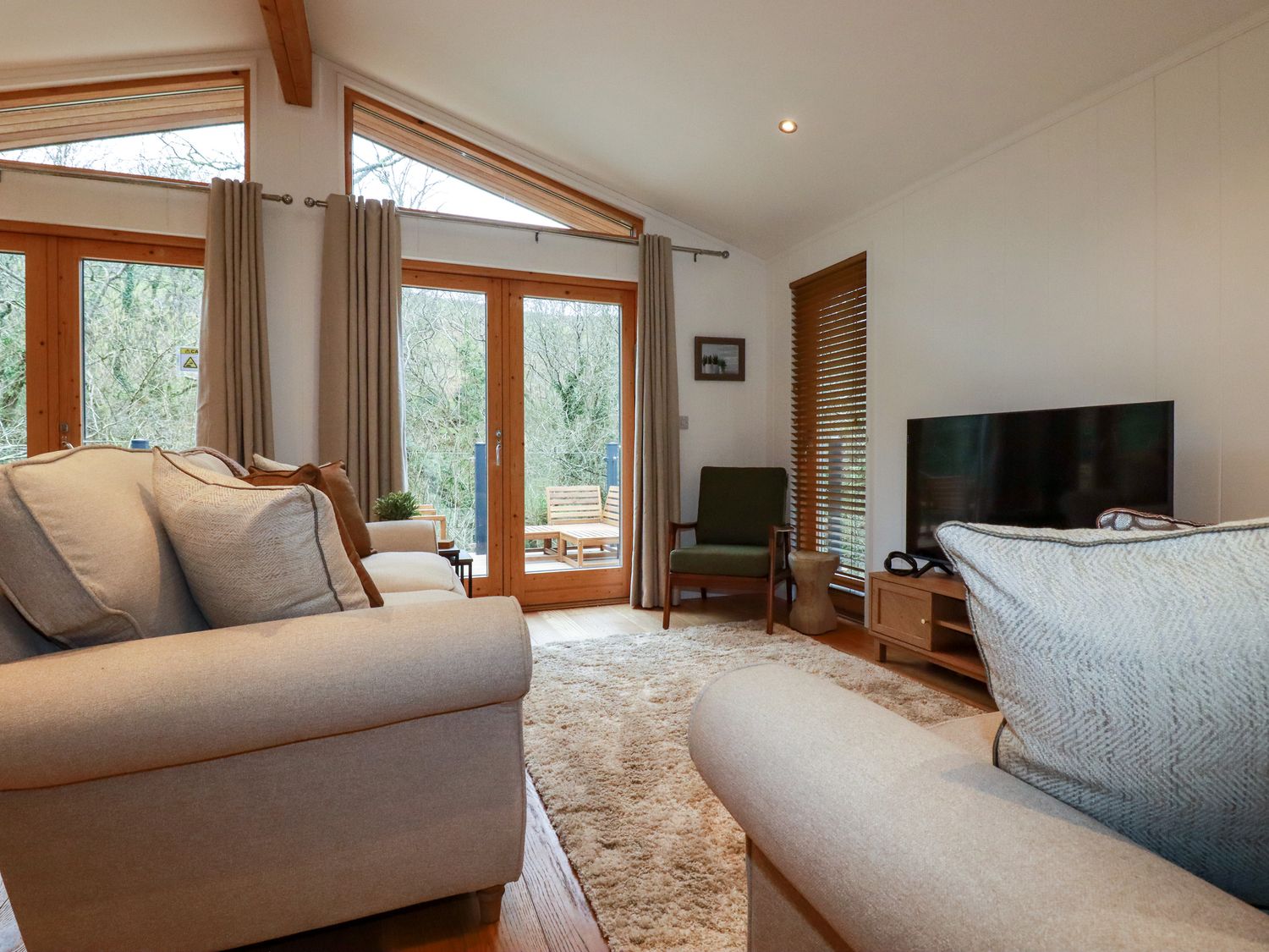 A living room with beige sofas a green armchair a television and wooden-framed windows at 1 Streamside in Lanreath