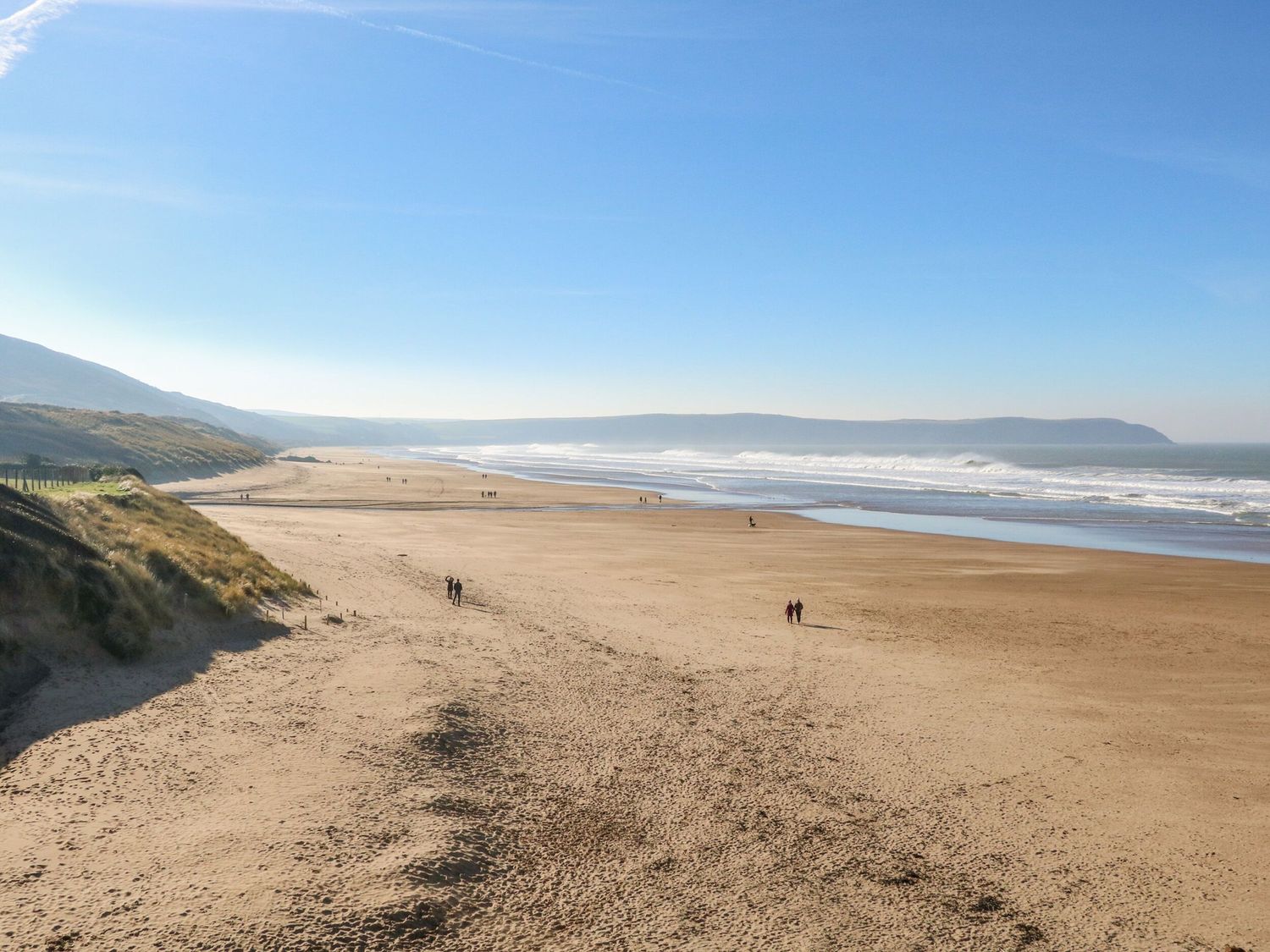 A wide sandy beach with several people walking near the shoreline and dunes on the left at Sunset Cottage in Swimbridge near Barnstaple