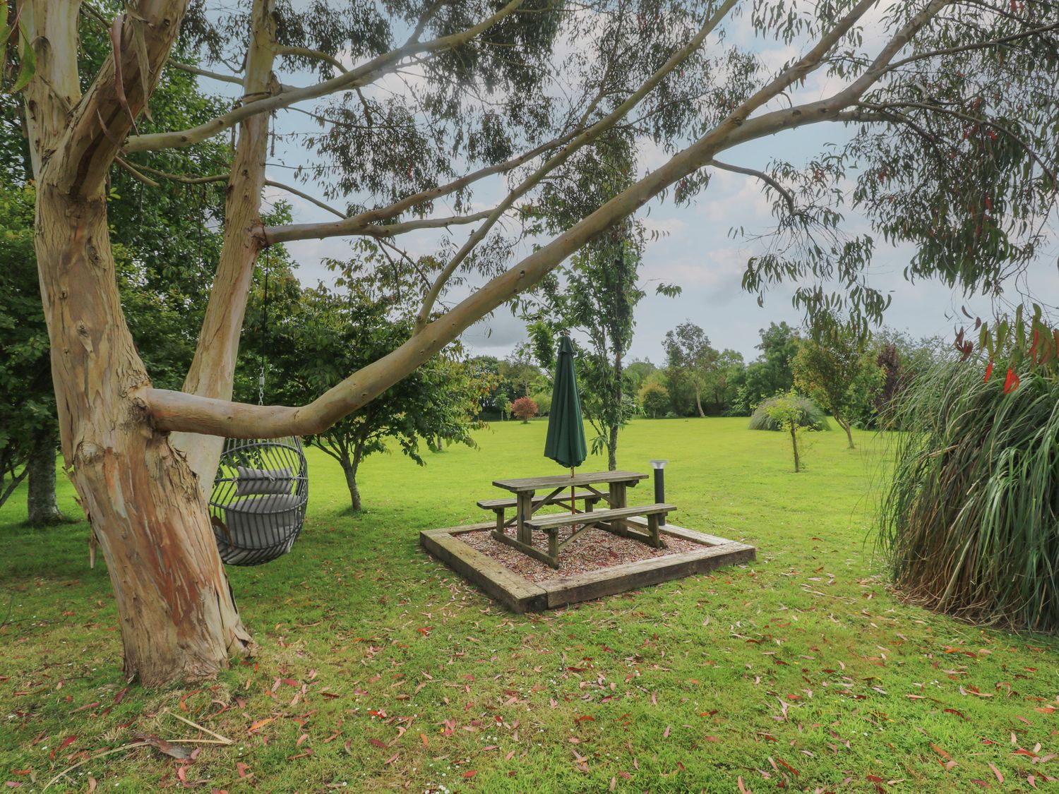 A garden with trees a wooden picnic table with an umbrella and a hanging chair from a tree at The Stables in Ruthvoes near St Columb Major