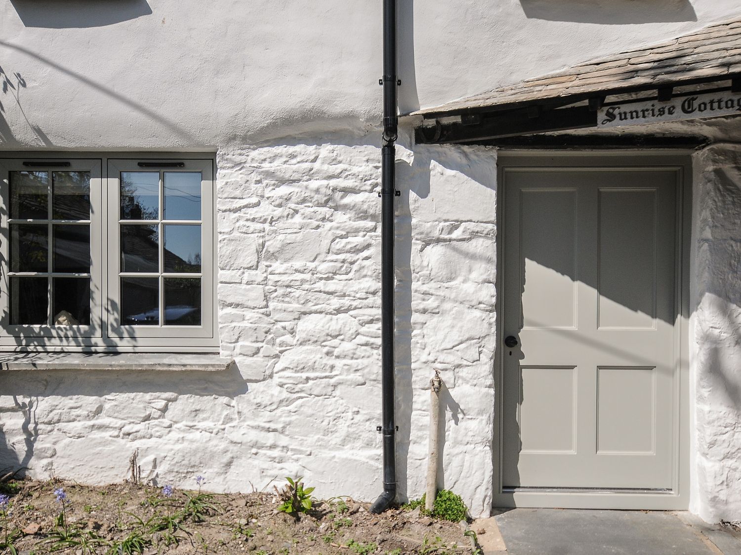 Exterior of a white stone cottage with a window and a door with a black drainpipe between them at Sunrise Cottage St Kew Highway near Wadebridge