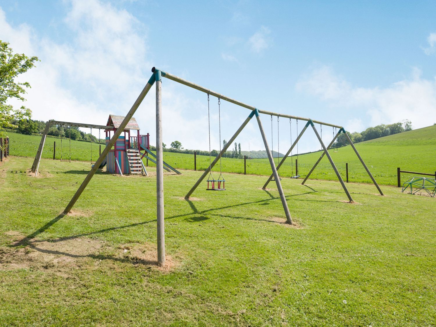 A playground with swings and a climbing structure in a grassy field at Owl Barn in Woolhope
