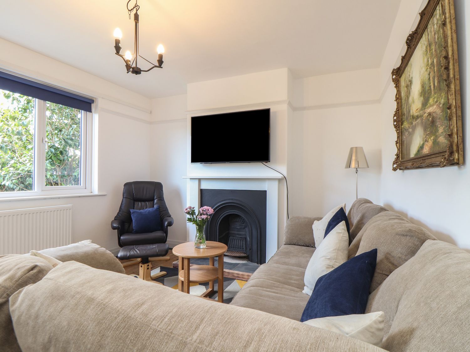 A living room with a beige sofa and blue cushions a black armchair a small round wooden table with flowers a wall-mounted TV and a fireplace at The Heights in Truro