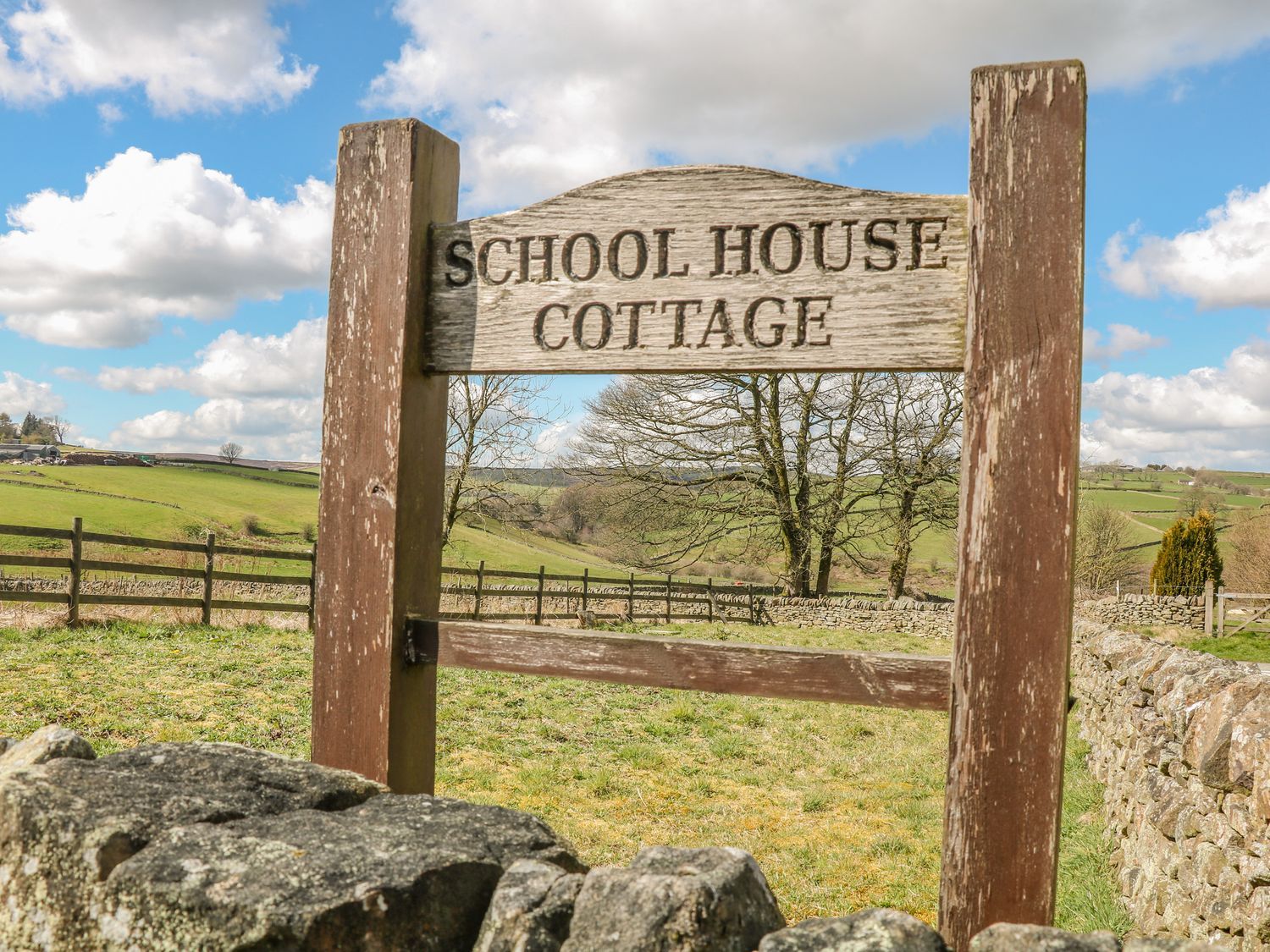A wooden sign reading School House Cottage with a rural landscape and stone fence in the background at School House Cottage in Newtown near Longnor